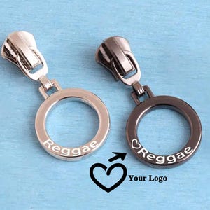 May include: Two metal zipper pulls with circular rings. The left pull is silver-toned and engraved with the word Reggae. The right pull is dark gunmetal-toned and engraved with a heart symbol and the word Reggae. Both feature a hinged attachment.