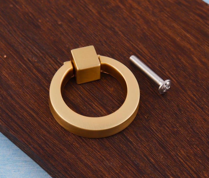 Op de afbeelding: Een goudkleurige kastgreep met een vierkante basis en een cirkelvormige handgreep. Naast de greep is een zilveren schroef geplaatst. De hardware is geplaatst op een donkerbruin houten oppervlak.