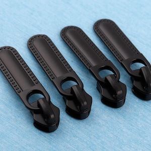 May include: Four matte black metal zipper pulls arranged in a diagonal row. Each pull features a long, slender, rounded rectangular tab with a decorative dotted border and a circular attachment ring connected to the main zipper slider mechanism.