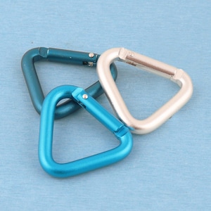 May include: Three triangular carabiners made of metal, featuring a spring-loaded gate mechanism. The set includes one dark teal carabiner, one bright blue carabiner, and one silver carabiner, all arranged in a cluster on a flat surface.
