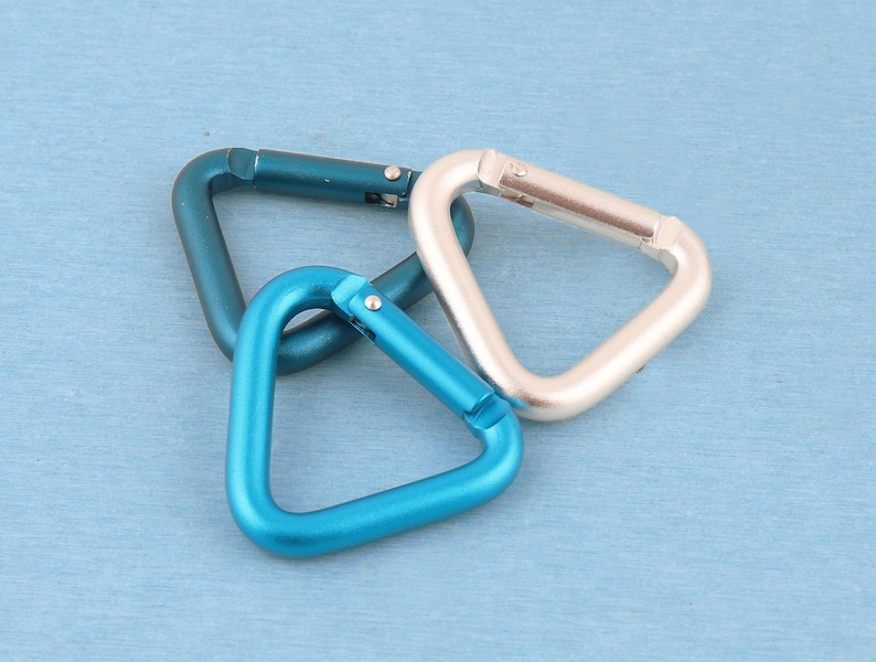 May include: Three triangular carabiners made of metal, featuring a spring-loaded gate mechanism. The set includes one dark teal carabiner, one bright blue carabiner, and one silver carabiner, all arranged in a cluster on a flat surface.