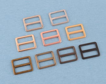 Op de afbeelding: Een verzameling van negen rechthoekige metalen gespen voor riemen, gerangschikt in drie rijen. De gespen zijn voorzien van een middenbalk en zijn verkrijgbaar in diverse metalen afwerkingen, waaronder zilver, goud, roségoud, brons, koper en matzwart.
