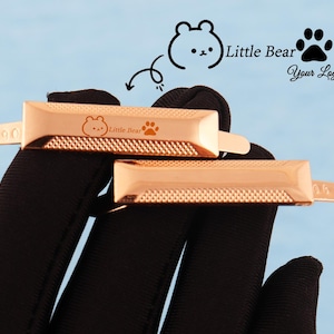 May include: Two rose gold metal clasps with a textured surface and rounded edges. Each clasp features a small bear graphic, the text "Little Bear", and a paw print. The clasps are displayed against a black background with a light blue backdrop.