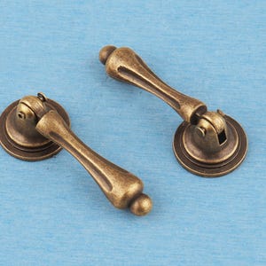 May include: Two antique brass-finished metal drop pulls, each featuring a circular mounting plate and a hinged, teardrop-shaped handle with a smooth, tapered design. The hardware has a weathered, darkened patina with visible highlights.