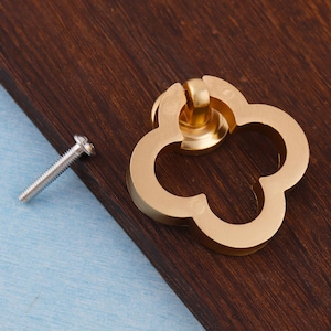 Może przedstawiać: Złoty uchwyt do szafki w kształcie koniczyny z pasującą śrubą. Okucia są umieszczone na ciemnobrązowej drewnianej powierzchni z jasnoniebieskim tłem. Uchwyt jest elementem dekoracyjnym mebli.