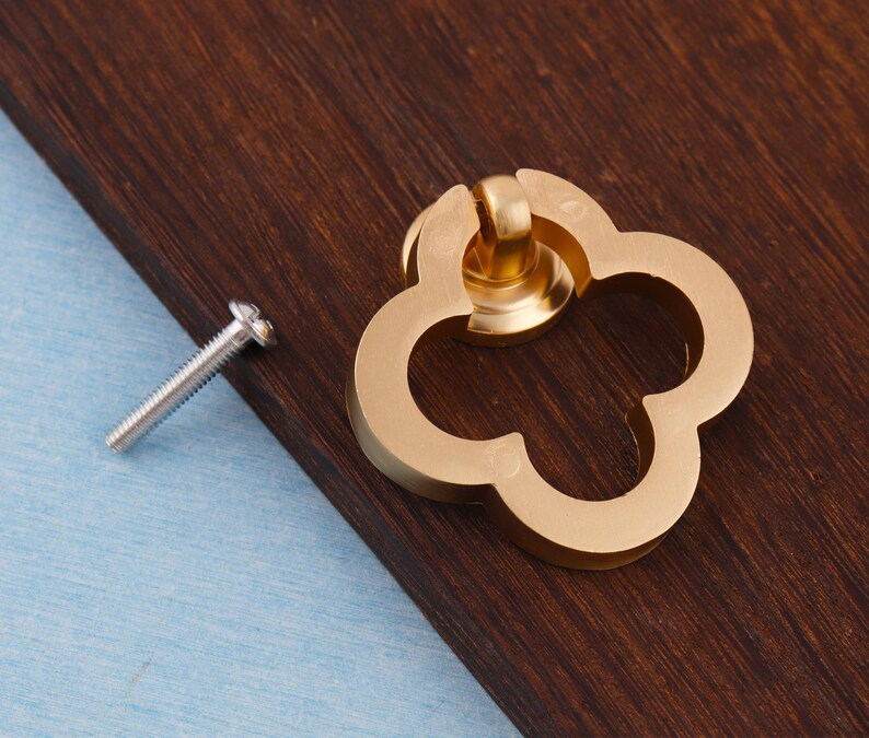 Może przedstawiać: Złoty uchwyt do szafki w kształcie koniczyny z pasującą śrubą. Okucia są umieszczone na ciemnobrązowej drewnianej powierzchni z jasnoniebieskim tłem. Uchwyt jest elementem dekoracyjnym mebli.