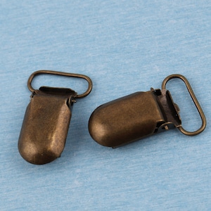 May include: Two antique gold-toned metal baby clothes clips. The clips have a rounded shape and a spring-loaded mechanism.