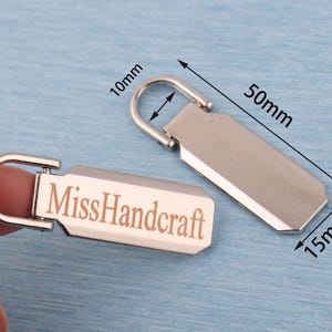 K&ouml;nnte beinhalten: Zwei silberfarbene Rei&szlig;verschluss-Anh&auml;nger mit rechteckigem Design. Einer der Anh&auml;nger tr&auml;gt den Schriftzug "MissHandcraft". Die Anh&auml;nger sind 5 cm lang, 1,5 cm breit und die Schlaufe ist 1 cm. Der Hintergrund ist hellblau.
