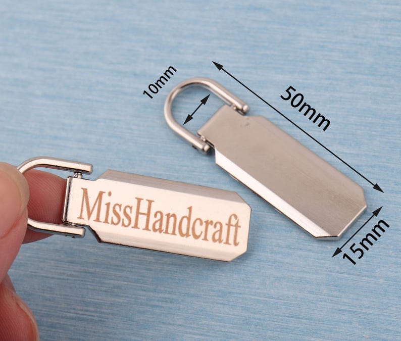 K&ouml;nnte beinhalten: Zwei silberfarbene Rei&szlig;verschluss-Anh&auml;nger mit rechteckigem Design. Einer der Anh&auml;nger tr&auml;gt den Schriftzug "MissHandcraft". Die Anh&auml;nger sind 5 cm lang, 1,5 cm breit und die Schlaufe ist 1 cm. Der Hintergrund ist hellblau.