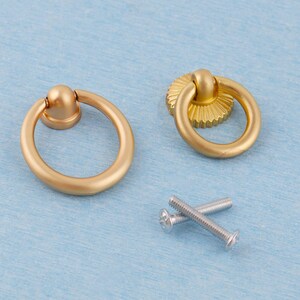 May include: Two gold-colored ring pull cabinet handles with a matte finish, and two silver screws. One handle has a simple round base, the other has a decorative, ridged base. The hardware is set against a light blue background.