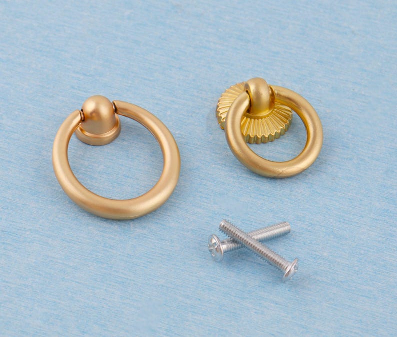 Op de afbeelding: Twee goudkleurige ringgreepkastknoppen met een matte afwerking en twee zilveren schroeven. E&eacute;n handvat heeft een eenvoudige ronde basis, de andere een decoratieve, gegroefde basis. De hardware staat tegen een lichtblauwe achtergrond.