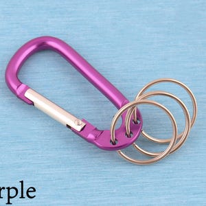 May include: A purple carabiner keychain featuring a spring-loaded silver gate and three attached circular metal key rings. The carabiner has a smooth, metallic finish and is designed with multiple holes at the base to hold the rings securely.