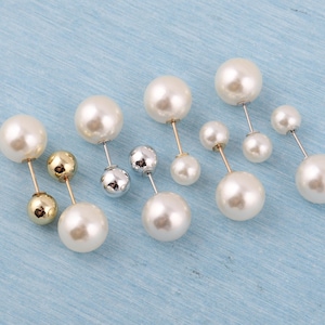 Puede incluir: Varias broches de perlas dispuestas sobre una superficie azul claro. Las broches presentan una perla grande, blanca y redonda combinada con una perla más pequeña. Algunas tienen postes dorados, otras plateados.