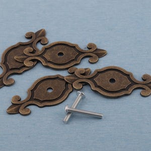 Könnte beinhalten: Zwei antike Schubladengriffe in Bronzefarbe mit kunstvollen Schnörkeln und einem ovalen Mittelteil. Zwei silberne Schrauben sind enthalten. Geeignet für Möbelrestaurierung oder Dekorationsprojekte.