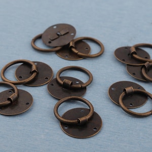 May include: Bronze-coloured drawer pulls with round handles and circular backplates. The hardware has a vintage aesthetic, with a slightly distressed finish. The pulls are designed for easy installation with pre-drilled holes.