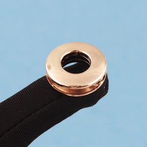 May include: A rose gold metal grommet or eyelet attached to a black fabric strap. The circular fastener features a polished, reflective surface with a central opening, securely crimped onto the textured, dark-colored textile material.