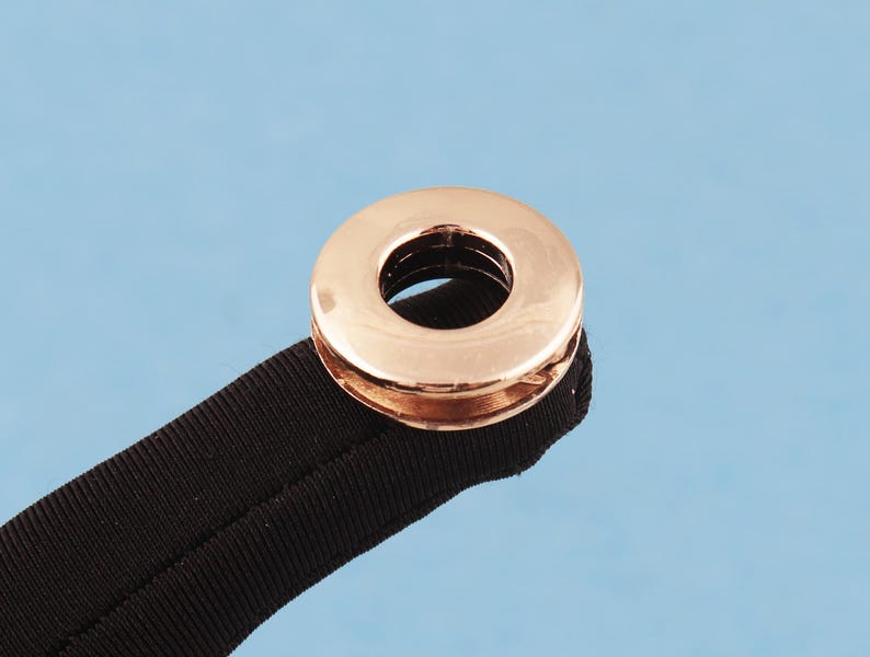 May include: A rose gold metal grommet or eyelet attached to a black fabric strap. The circular fastener features a polished, reflective surface with a central opening, securely crimped onto the textured, dark-colored textile material.