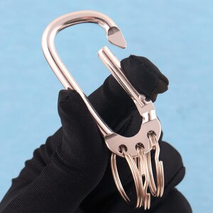 May include: A rose gold-toned metal carabiner keychain featuring a spring-loaded gate mechanism and a semi-circular base with four integrated small circular rings attached for holding keys or other small items securely in a compact arrangement.
