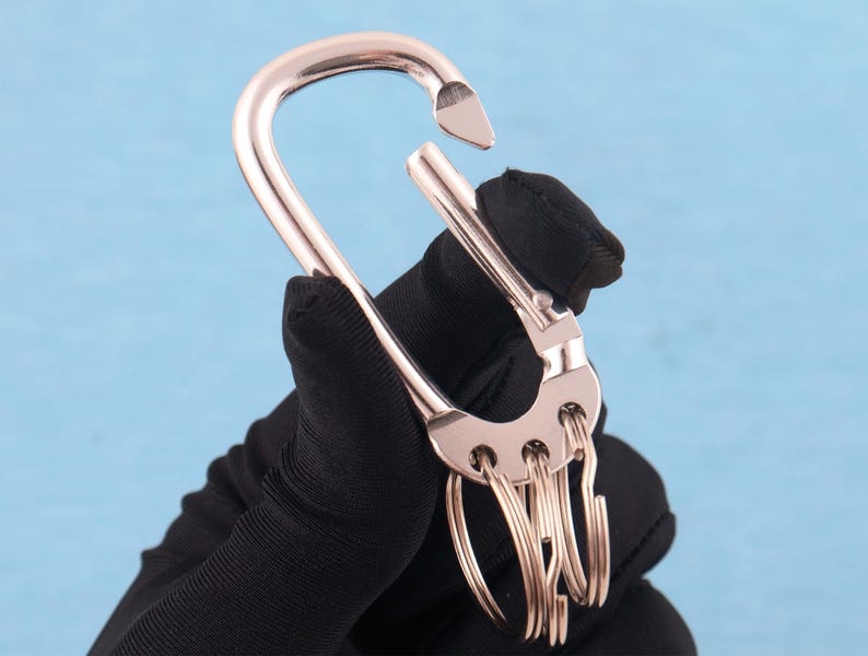 May include: A rose gold-toned metal carabiner keychain featuring a spring-loaded gate mechanism and a semi-circular base with four integrated small circular rings attached for holding keys or other small items securely in a compact arrangement.