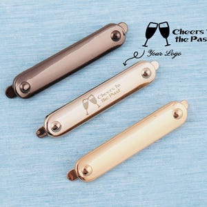 May include: Three metallic keychains in bronze, rose gold, and gold, each with a rounded rectangular shape. The middle keychain has the text "Cheers to the Past" and two champagne glasses. The background is a light blue surface.