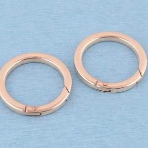 Puede incluir: Dos anillas circulares de resorte hechas de metal en tono oro rosa. Cada anilla presenta un acabado liso y pulido, con un pequeño mecanismo de resorte integrado que permite abrir y cerrar la anilla para sujetar diversos objetos.