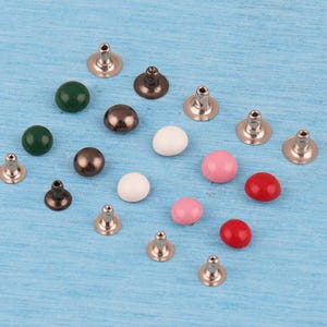 May include: A collection of small, round metal rivets with smooth, colorful domed caps in shades of dark green, metallic brown, white, light pink, and bright red, each featuring a silver-toned metal post on the reverse side for attachment.