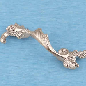 May include: A metallic silver-toned cabinet pull featuring an elongated, curved shape with intricate leaf-like textures and detailing. The handle has two circular mounting points at each end, designed for attachment to furniture or cabinetry doors.