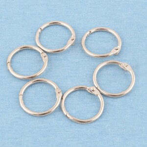 May include: A set of six circular silver-toned metal binder rings arranged in a loose cluster. Each ring features a hinged opening mechanism with a small rivet, allowing the ring to be opened and closed for securing documents or various items.
