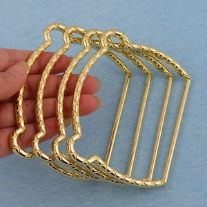 May include: A collection of gold-colored metal hair combs. Each comb has a decorative, textured design along the curved top and straight, parallel tines. The combs are held in a hand against a light blue background.