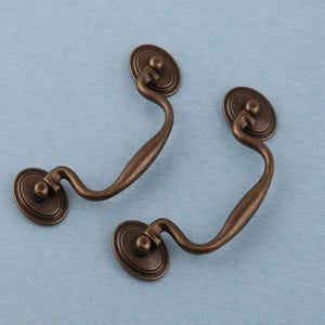 May include: Two antique-style bronze drawer pulls. Each pull has an oval backplate and a curved handle. The hardware has a distressed finish, suggesting an aged appearance. Suitable for furniture restoration or adding vintage charm.