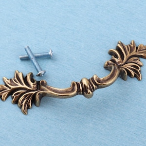 May include: An antique brass-toned metal drawer pull featuring an ornate, leaf-inspired design with curved edges and intricate detailing. The handle is accompanied by two silver-coloured metal mounting screws with threaded shafts and flat heads.