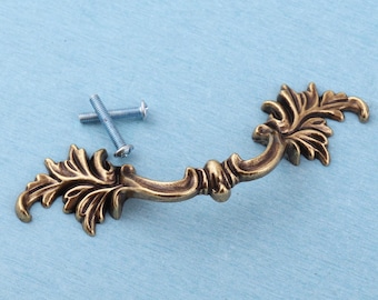 May include: An antique brass-toned metal drawer pull featuring an ornate, leaf-inspired design with curved edges and intricate detailing. The handle is accompanied by two silver-colored metal mounting screws with threaded shafts and flat heads.