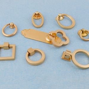 Könnte beinhalten: Eine Sammlung goldfarbener Schrankbeschläge, darunter Ringgriffe, quadratische Griffe und ein dekorativer Griff mit floralem Design. Die Beschläge sind auf einer hellblauen Oberfläche angeordnet und zeigen verschiedene Formen und Stile.