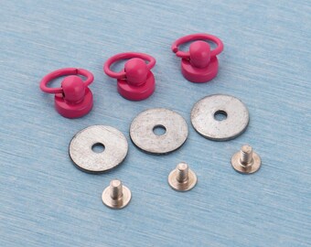 May include: A collection of pink metal D-ring rivets with matching washers and screws. The rivets have a circular ring attached to a rounded base. The washers are silver and round with a central hole. The screws are silver.