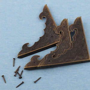 May include: Bronze-coloured corner brackets with intricate scrollwork and small nails. The triangular brackets are designed for corner reinforcement. Ideal for woodworking projects, these brackets add a vintage aesthetic to furniture or boxes.