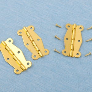 May include: Three gold-colored decorative hinges with matching screws are arranged on a light blue surface. Each hinge has a decorative shape with rounded edges and multiple screw holes. The hinges are made of metal and have a shiny finish.