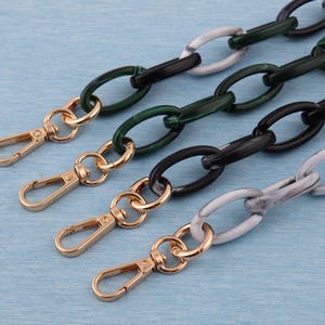 May include: Close-up of four handbag chains with gold-colored clasps. The chains feature oval links in black, green, and marbled gray and white. The chains are arranged diagonally against a light blue background.