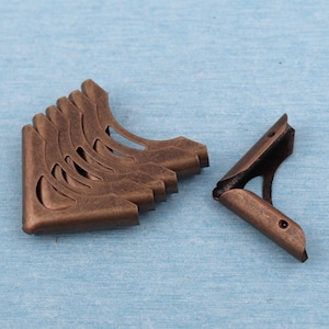 Könnte beinhalten: Eine Sammlung antiker Eckenschützer aus Metall in Bronzeoptik. Die dekorativen Eckwinkel haben ein geschwungenes Design mit ausgeschnittenen Details. Ein Eckwinkel ist vom Stapel getrennt. Wahrscheinlich für Bastelarbeiten oder Buchbinderei.