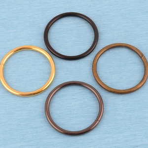 May include: Four circular metal rings arranged in a cluster. The set includes one polished gold-tone ring, one matte black ring, one dark bronze-tone ring, and one copper-tone ring. Each ring features a smooth, continuous surface with a visible seam.