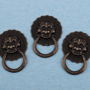 Può includere: Tre battenti antichi in bronzo con un design a testa di leone. Ogni battente presenta una faccia circolare con una testa di leone dettagliata e una maniglia ad anello. I battenti sono su uno sfondo azzurro.