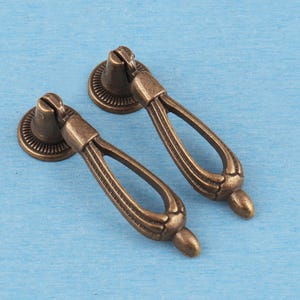 Peut inclure: Deux poignées de tiroir en métal de couleur bronze antique, dotées d'une poignée pivotante en forme de goutte d'eau fixée sur une base de montage circulaire et nervurée. La quincaillerie présente une finition texturée et patinée.