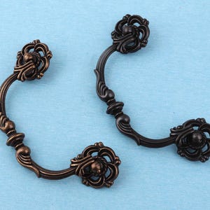 May include: Two curved metal drawer pulls with a dark bronze finish. Each handle features an ornate, swirling floral design at both ends, with a central decorative element on the arched bar connecting the two circular mounting plates.