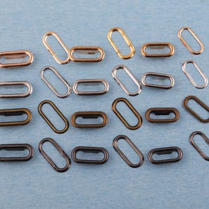 Puede incluir: Un conjunto de ojales metálicos ovalados en varios acabados, incluyendo dorado, plateado, bronce y negro. Estos componentes de ferretería están dispuestos sobre una superficie azul claro, mostrando sus diferentes colores y texturas. Ideal para proyectos de artesanía y costura.