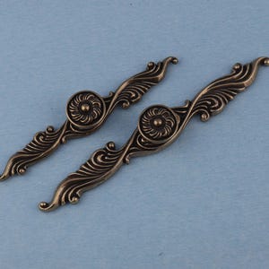 May include: A pair of antique bronze-coloured drawer pulls, each featuring a floral centre and ornate, curved edges. The hardware is set against a light blue background, highlighting the decorative details.