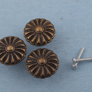 Könnte beinhalten: Drei bronzefarbene Schrankknöpfe in Blumenform mit einem zentralen kreisförmigen Design und strahlenförmigen Blütenblättern. Zwei silberne Schrauben sind ebenfalls vorhanden. Die Knöpfe befinden sich auf einer hellblauen Oberfläche.