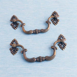 Puede incluir: Dos tiradores de cajón antiguos de bronce con detalles florales ornamentados. Los tiradores tienen un diseño de asa curvada y están colocados sobre un fondo azul claro. Estas piezas de ferretería decorativas son ideales para proyectos de restauración de muebles.