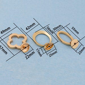 Może przedstawiać: Trzy złote uchwyty do szafek o r&oacute;żnych kształtach: czterolistna koniczyna (57 mm x 49 mm), koło (56 mm x 42 mm) i owal (55 mm x 30 mm). Każdy uchwyt ma podstawę montażową.