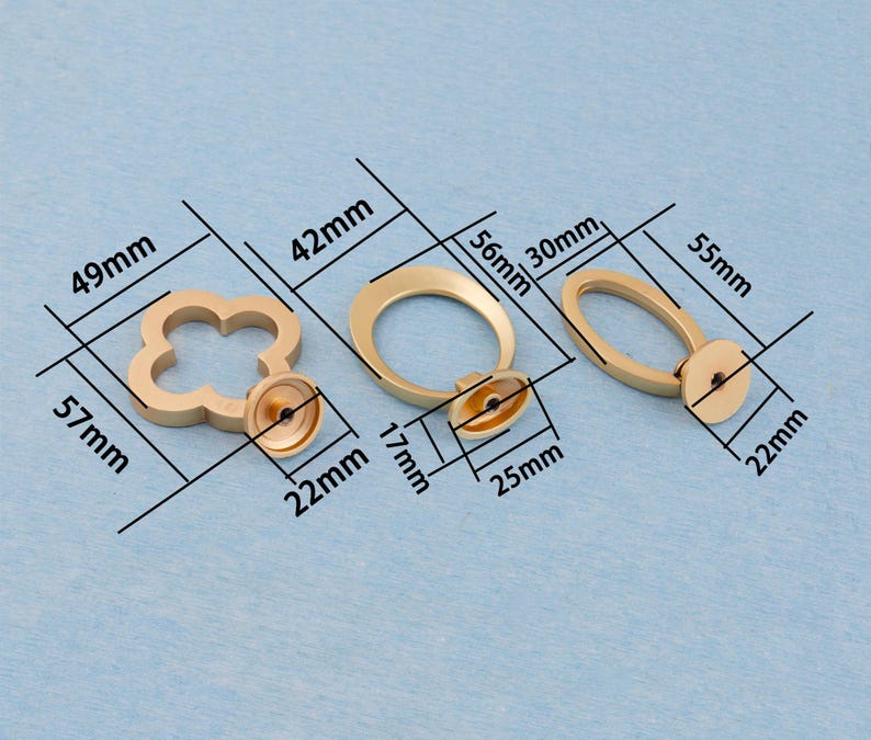 Może przedstawiać: Trzy złote uchwyty do szafek o r&oacute;żnych kształtach: czterolistna koniczyna (57 mm x 49 mm), koło (56 mm x 42 mm) i owal (55 mm x 30 mm). Każdy uchwyt ma podstawę montażową.