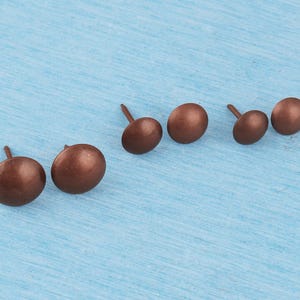Puede incluir: Un conjunto de seis pequeñas tachuelas redondas para tapicería con un acabado mate en tono cobre. Cada tachuela presenta una cabeza abovedada y un clavo metálico corto y puntiagudo en la base, dispuestas sobre una superficie azul claro.