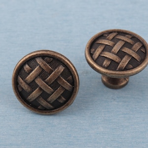 Pode incluir: Dois puxadores de armário em bronze com um design de cesta tecida. Os puxadores têm formato redondo com textura detalhada e uma haste pequena para montagem. O fundo é azul claro.